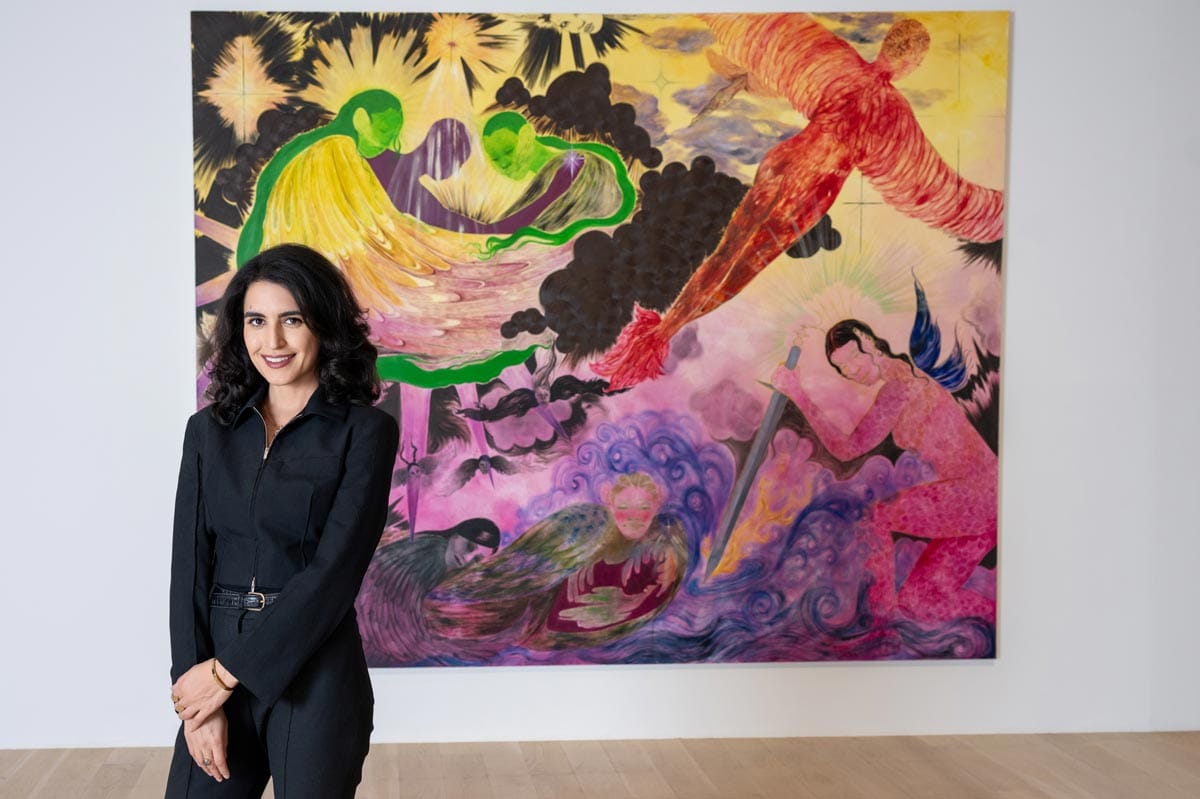 Donna stands in front of colorful painting with forms in pinks, purples, and black.