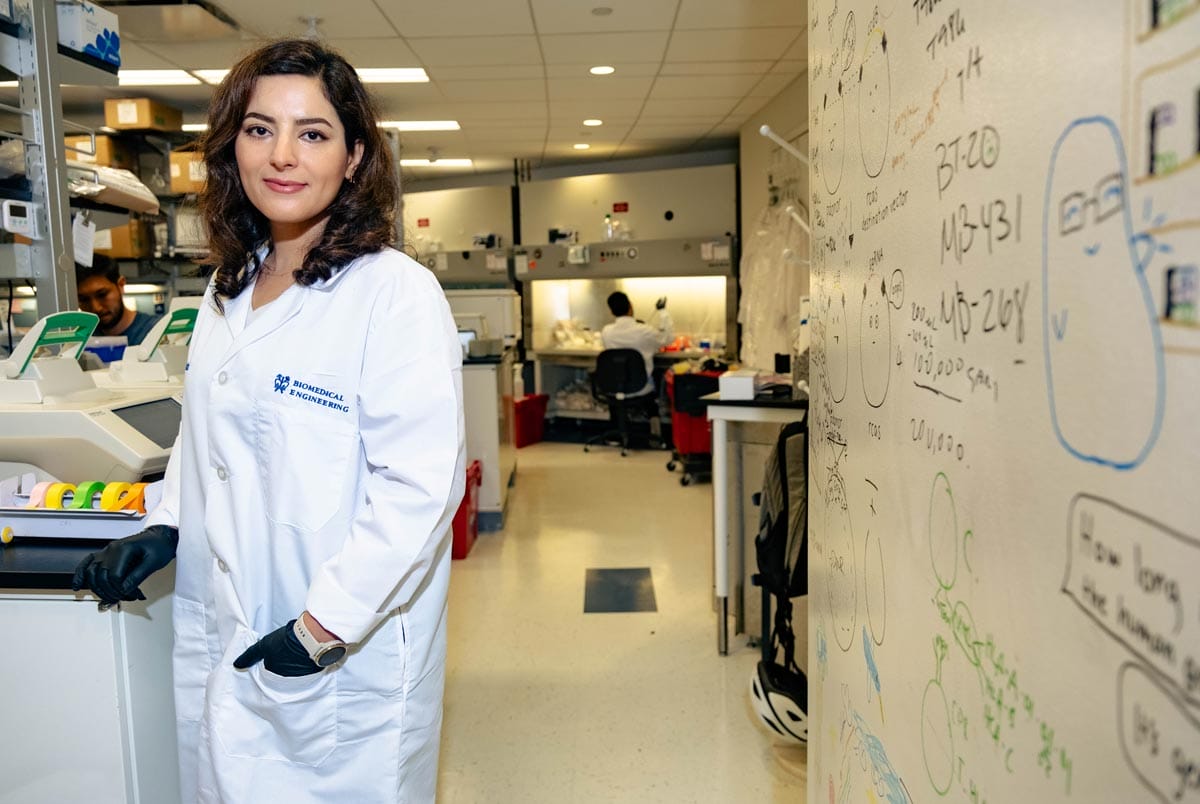 Elham standing next to a white board in her lab amongst machinery and equipment.