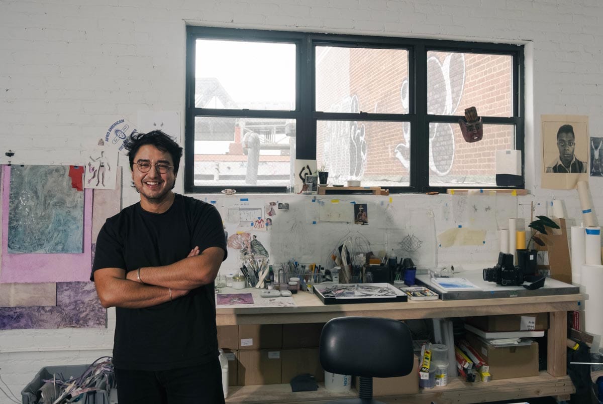 Felipe crosses his arms and smiles as he stands to the left of the large window in his studio.