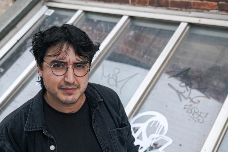 Felipe wearing glasses with round lenses and standing in front of an exterior skylight.