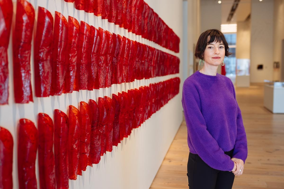 Francesca stands to the right of a wall with lots of small red art objects pinned to it.