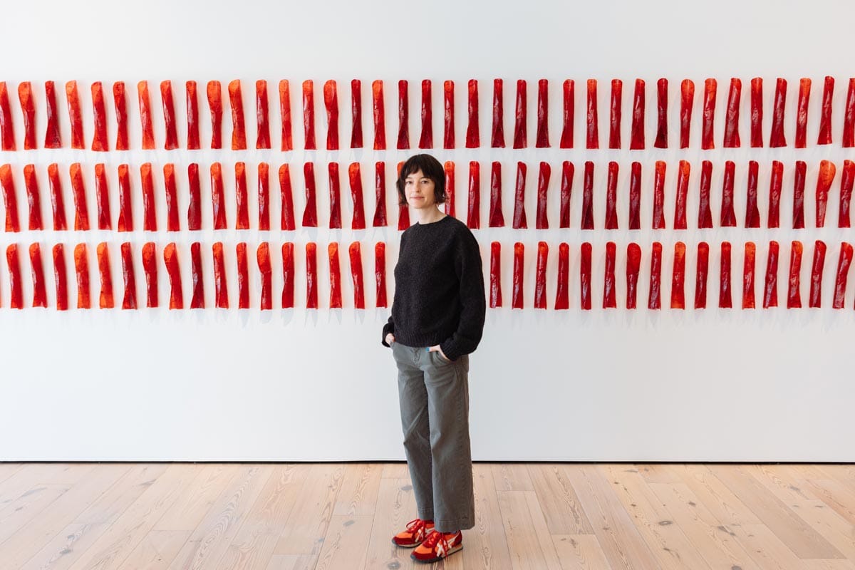 Francesca stands in a gallery space in front of a wall pinned with red art objects.