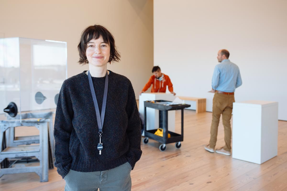 Francesca standing in a gallery with her colleagues working behind her her during an exhibition installation.