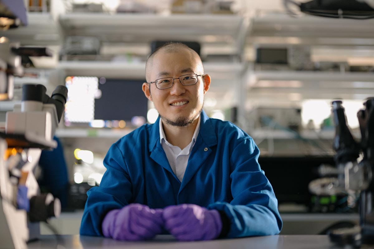 Guosong sitting in his lab, wearing a blue lab coat and purple gloves.