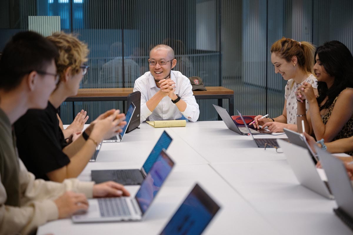 Guosong seated at the head of a conference table working with his lab members who are also seated at the table with their laptops open.