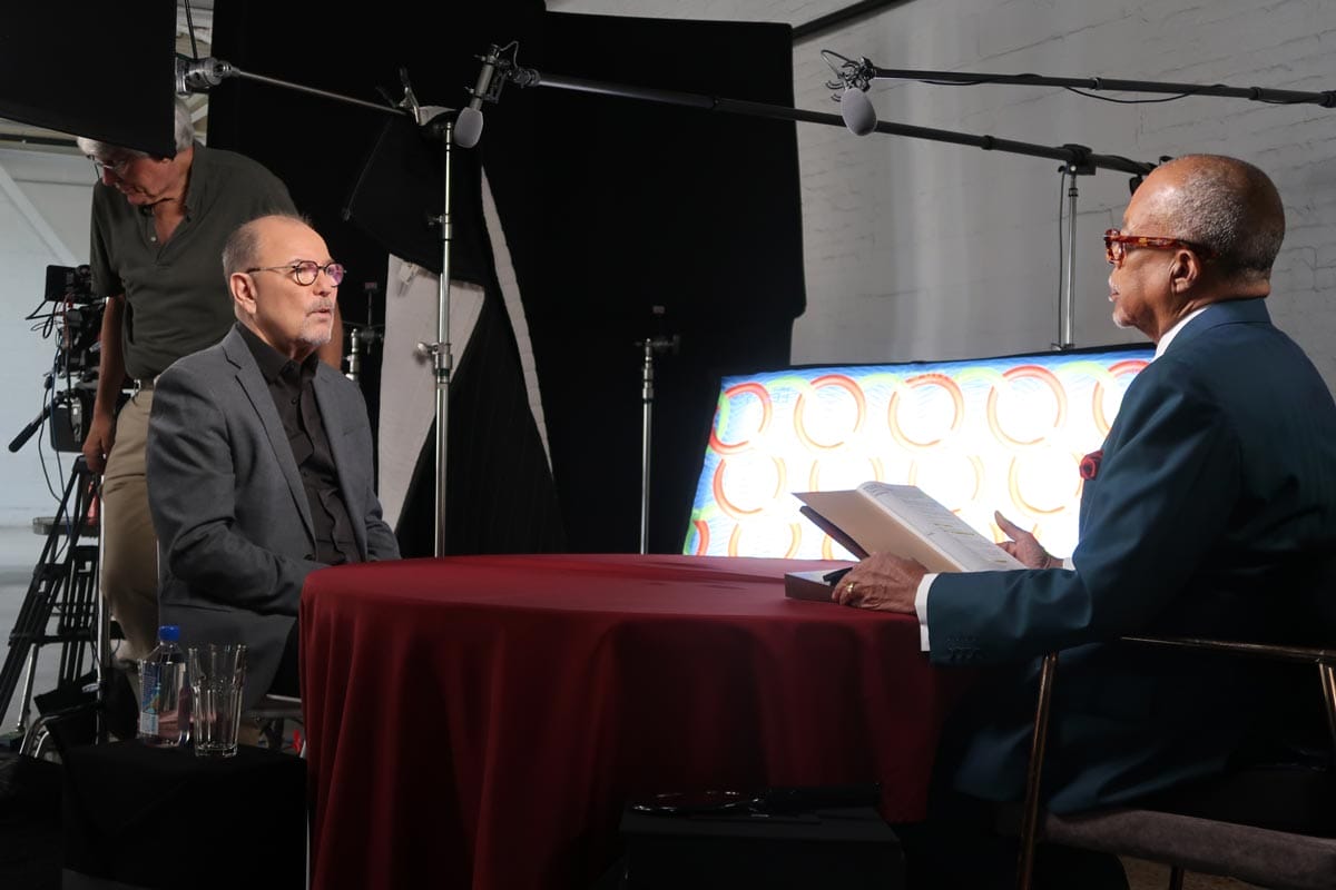 Henry Louis Gates, Jr. and Ruben Blades sit across from each other at a table with a burgundy tablecloth. 