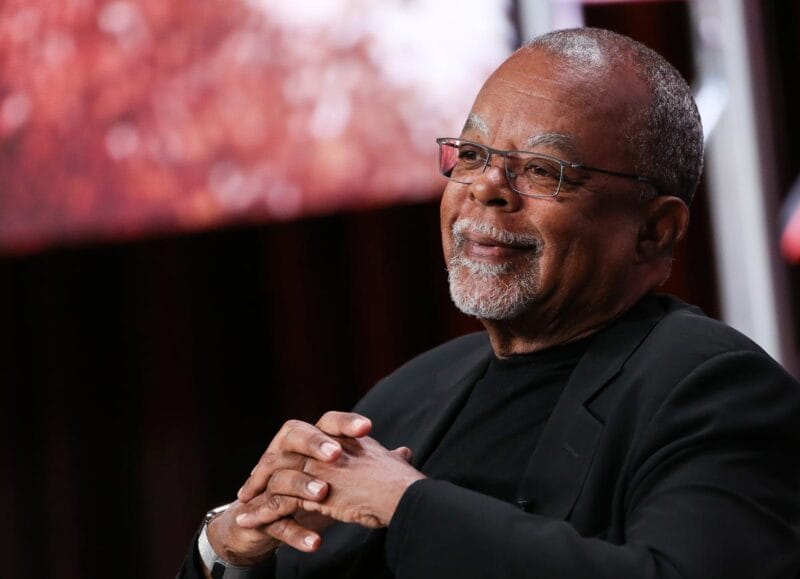 Henry Louis Gates Jr. sitting with his hands clasped in front of a red screen.