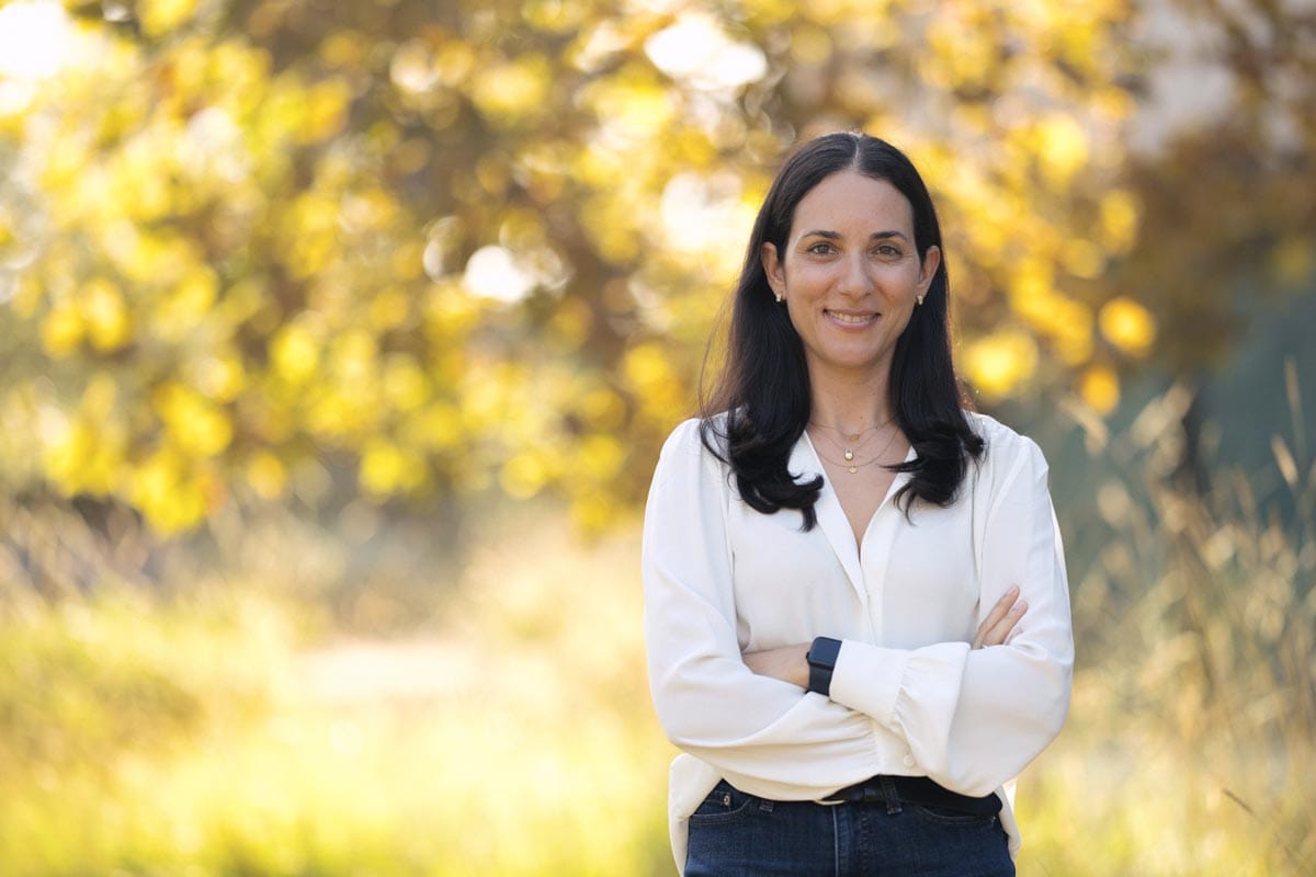 Maayan crosses her arms as she stands in front of branches with yellow leaves.