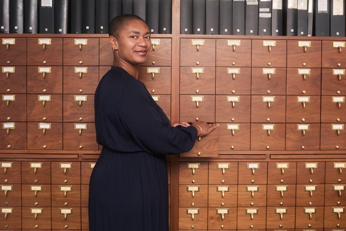 Oluremi standing in front a wall of small, wooden archival drawers.