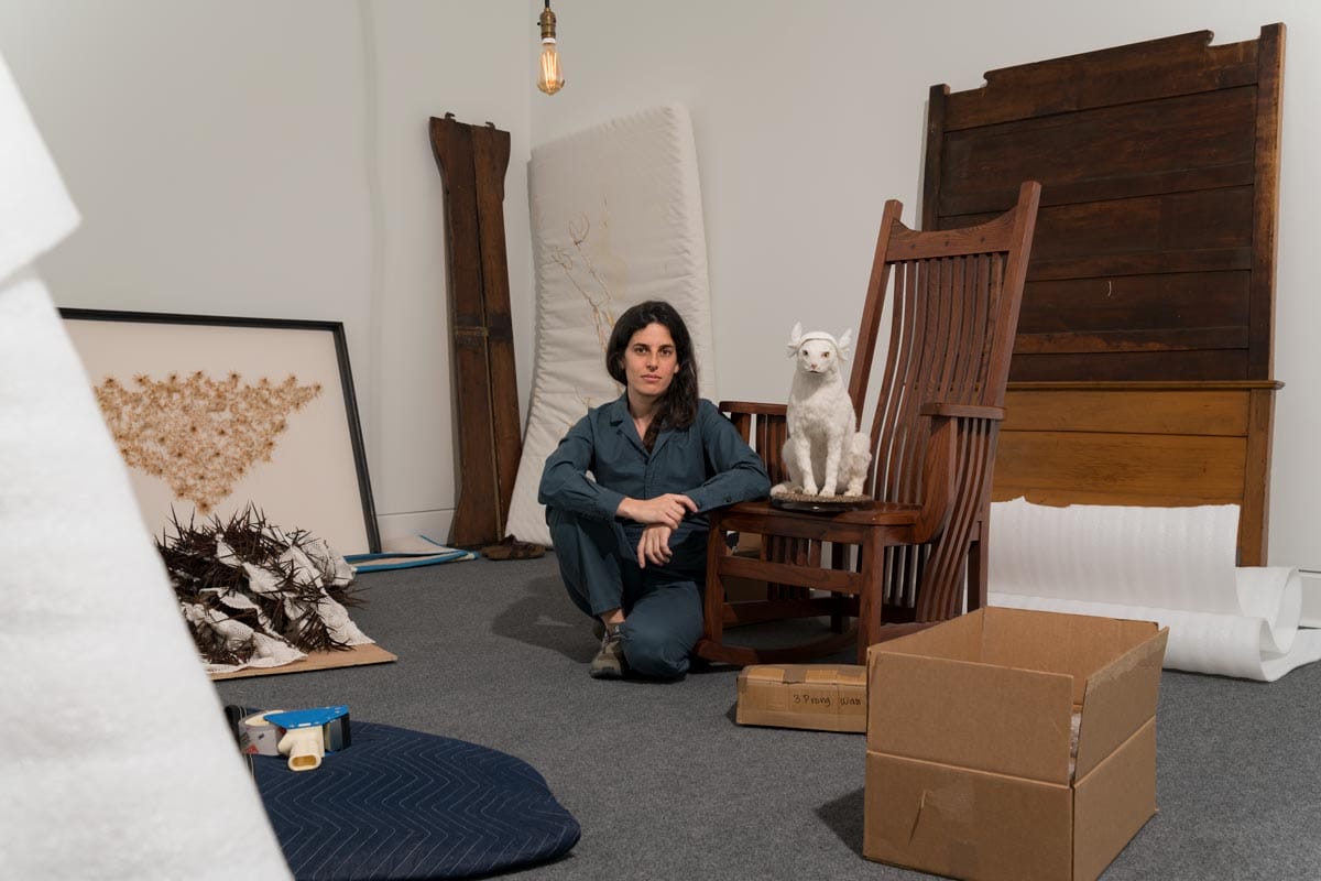 Selva seated in a room surrounded by her artworks - including a wooden chair and a taxidermy white cat.