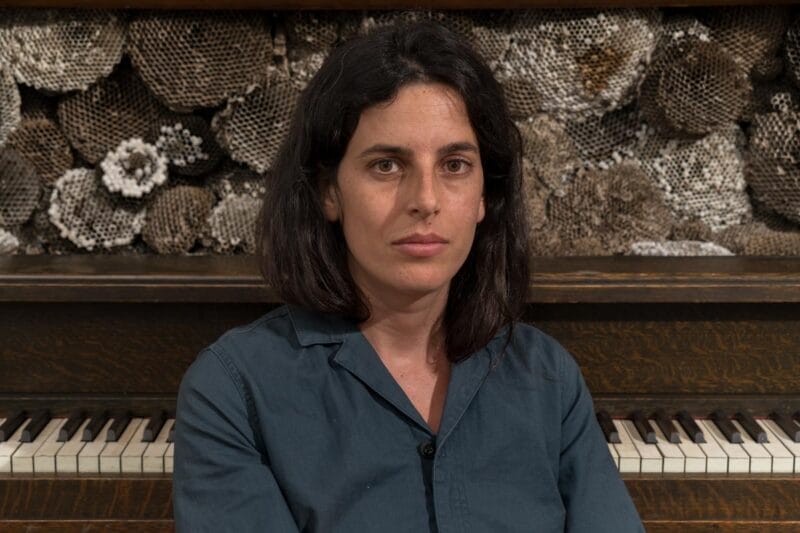 A close shot of Selva sitting at a piano with bee hives behind her.