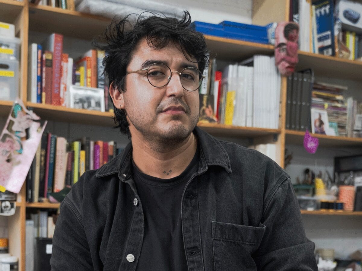 A portrait of Felipe Baeza with a full and busy bookshelf behind him. 