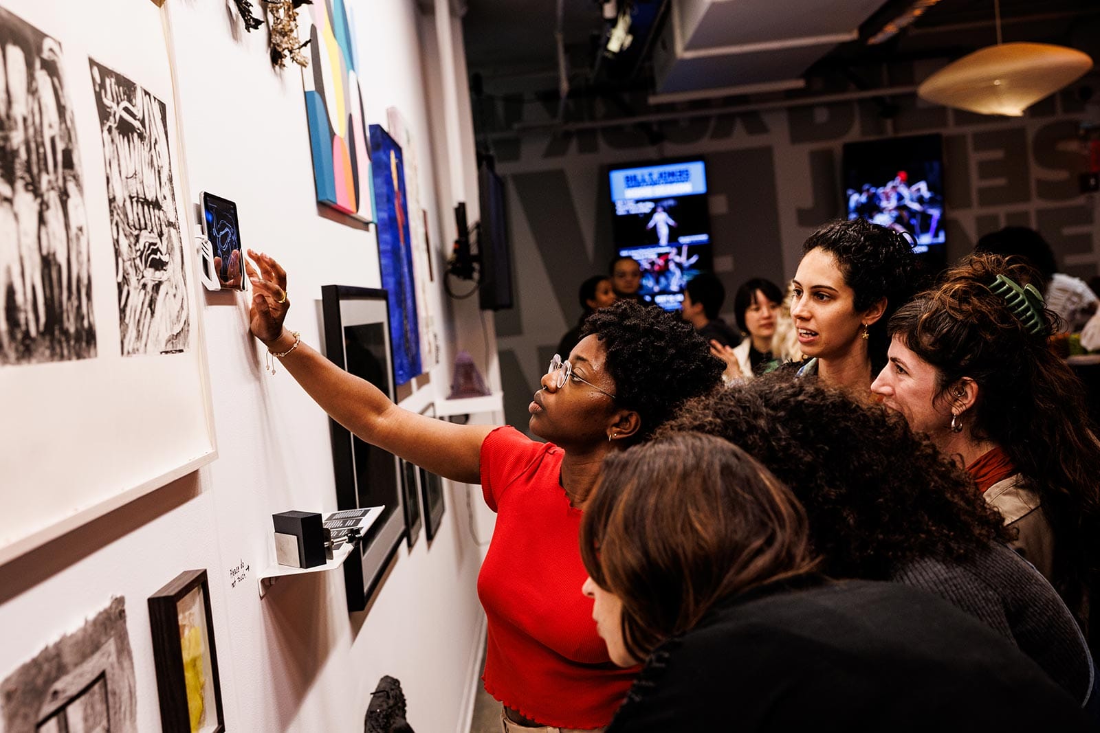 A woman in a red shirt reaches up to interact with an exhibit on a white wall, while other people gather around her looking at the artwork.