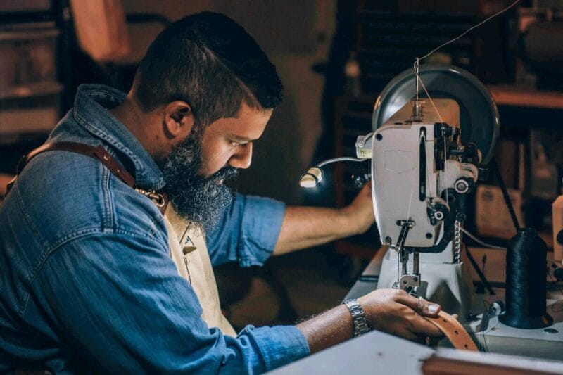 The side profile of a bearded man wearing denim as he uses a sewing machine to construct a leather belt.