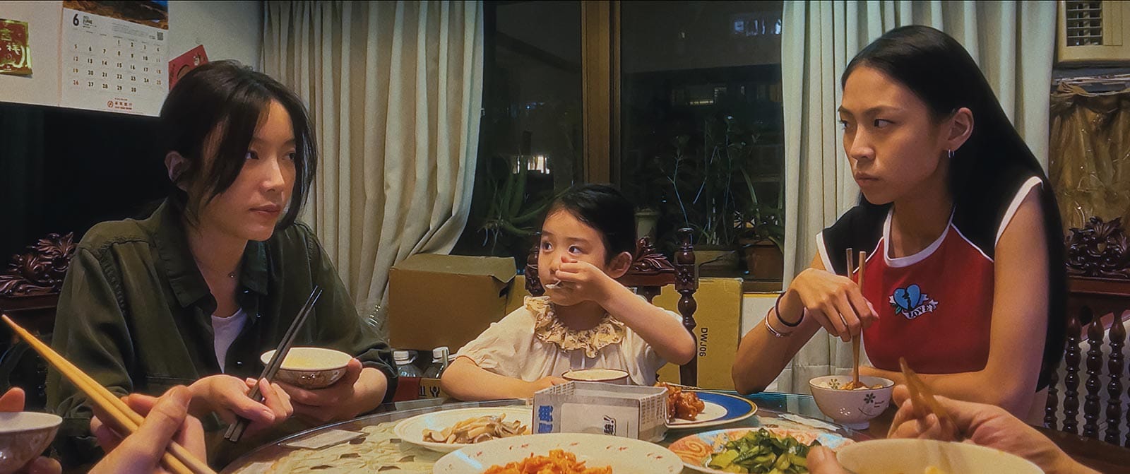 Three people sit at a dinner table, eating in tense silence. A woman and a teenage girl face each other while a young girl sits between them, glancing nervously as food fills the table.