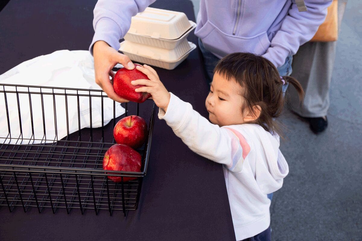 A toddler reaches for a red apple in a metal basket on a table.