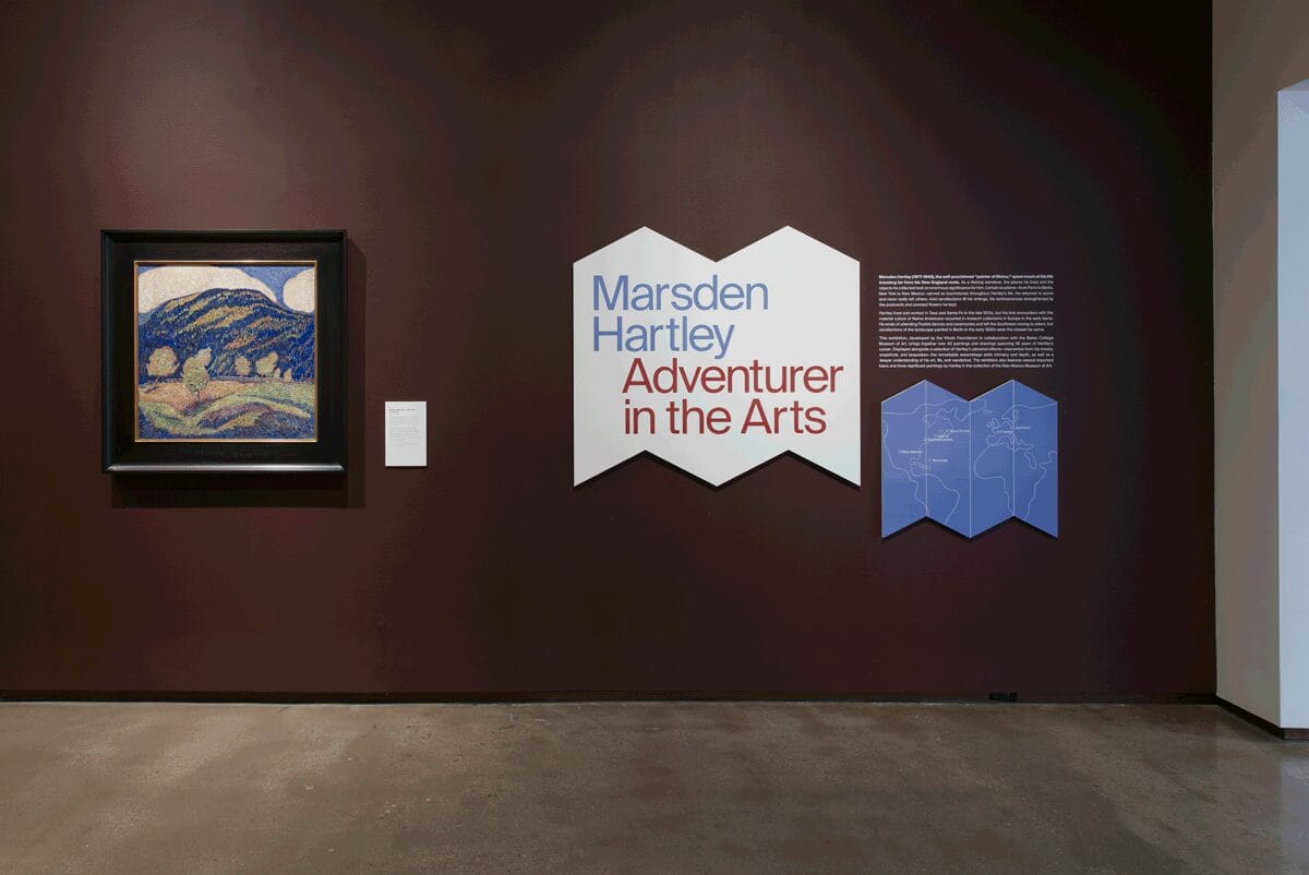 Marsden Hartley exhibition still: (left to right) Silence of Noon - Midsummer hangs on a burgundy wall next to a white cutout with "Marsden Hartley Adventurer in the Arts" in blue and red text.