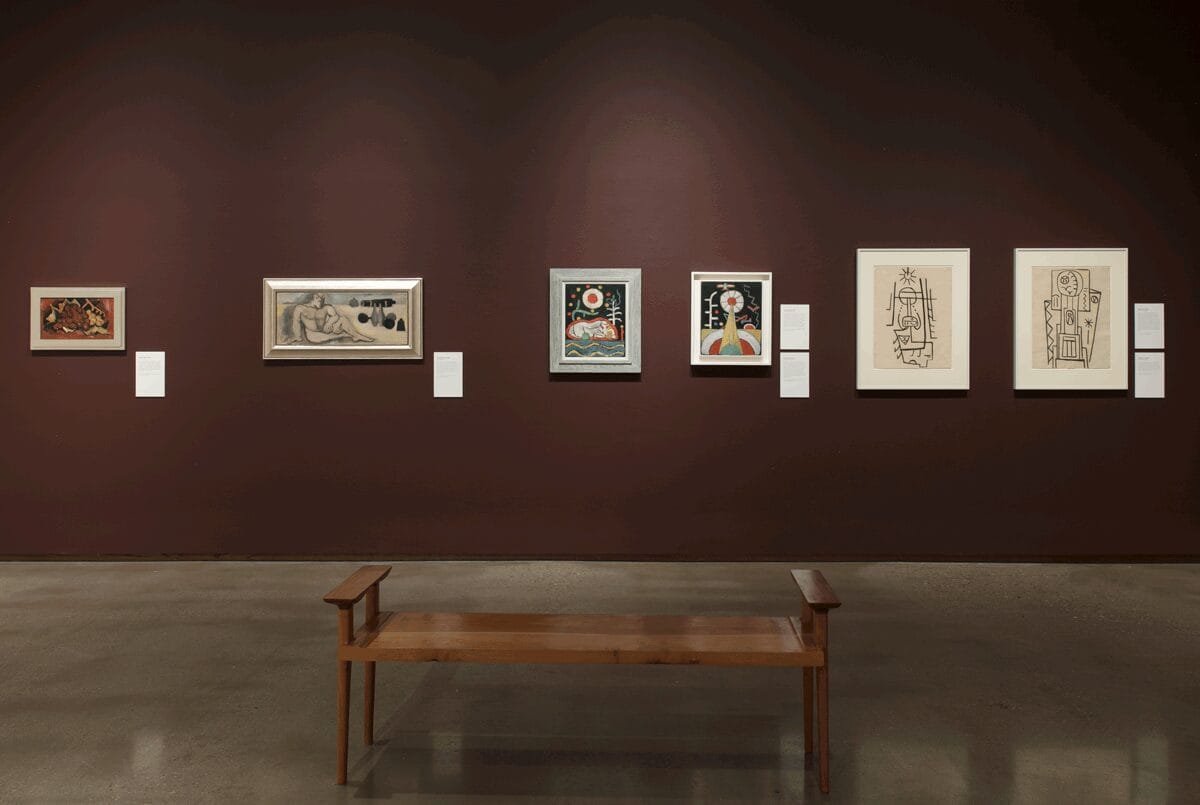 Marsden Hartley exhibition still: A burgundy wall with six of Hartley's paintings and a wooden bench centered on the concrete floor in front of them.