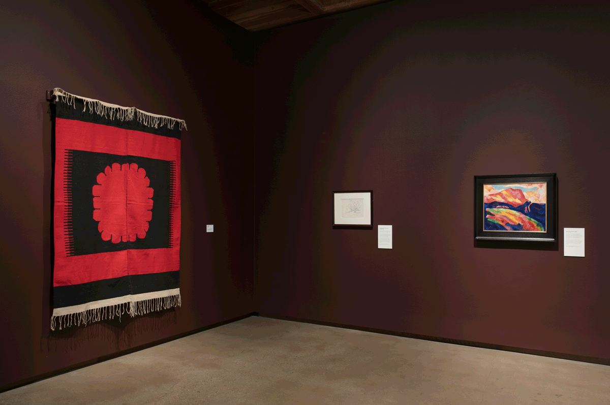 Marsden Hartley exhibition still: A red-and-black carpet with a large center medallion in the shape of a flower hangs on the left wall, and there are two works on the right wall: A drawing and Hartley's Mont Sainte-Victoire painting.