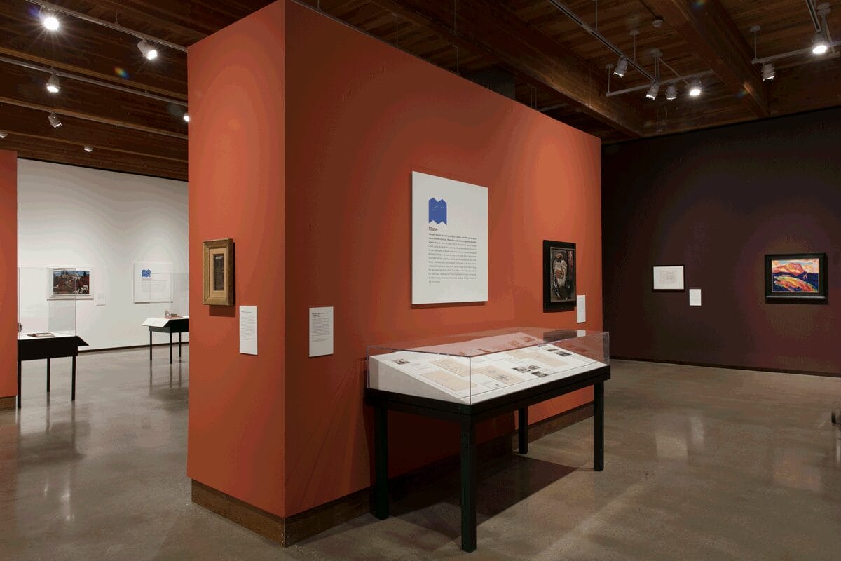 Marsden Hartley exhibition still: paintings are hung on white, burgundy, and burnt orange walls, and there is a display case with personal artifacts.