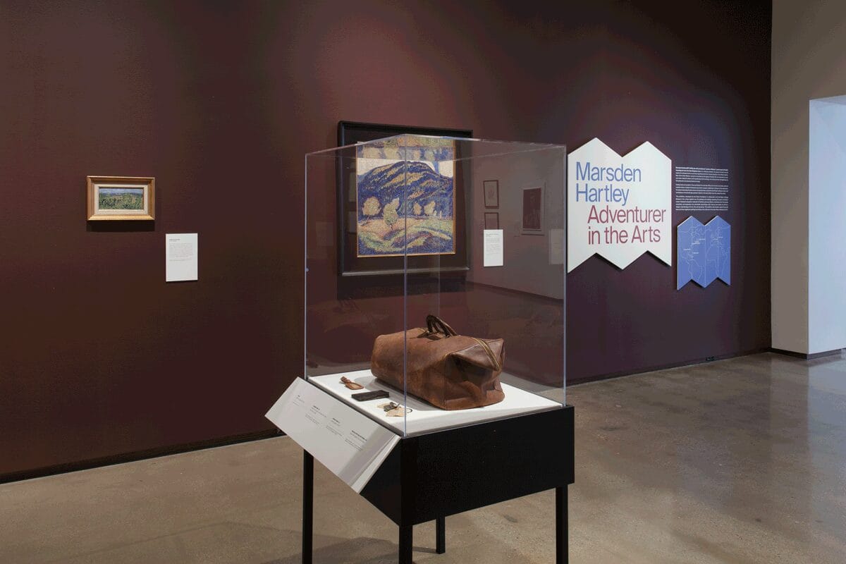 Marsden Hartley exhibition still: A display case with a travel bag and behind it Silence of Noon - Midsummer hangs on a burgundy wall next to a white cutout with
