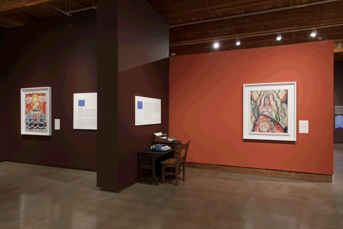 Marsden Hartley exhibition still: (left to right) Schiff displayed on a burgundy wall, a desk with a type-writer, and Portrait Arrangement No.2 displayed on a burnt orange wall.