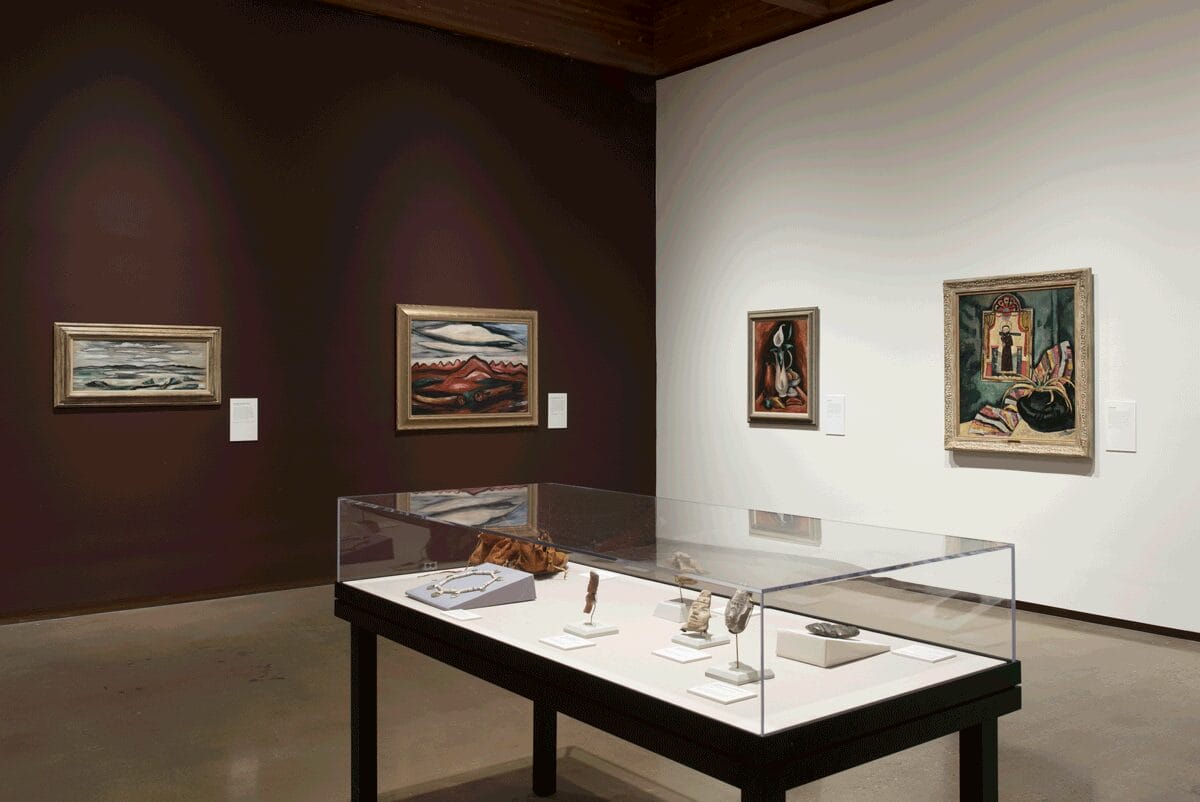 Marsden Hartley exhibition still: Two paintings on a burgundy wall, two paintings on a white wall, and a display case with his personal items.