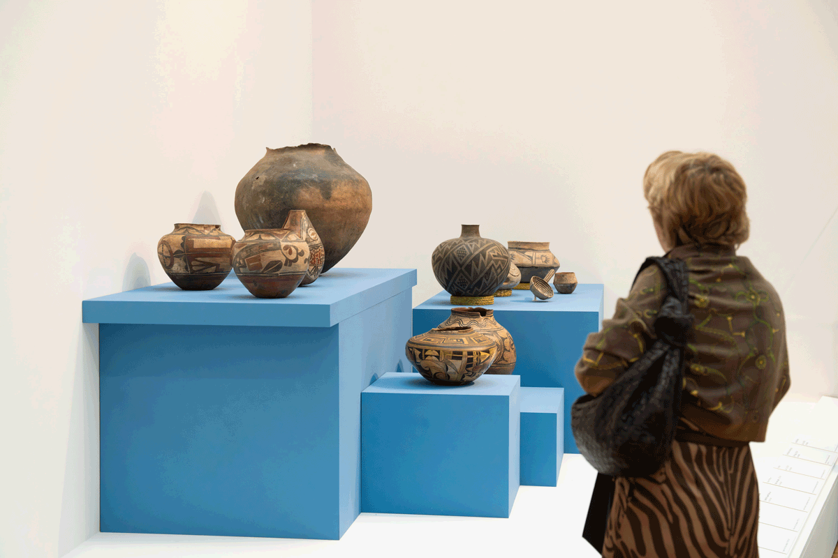 A person looking at pieces of pottery placed upon light blue pedestals of varying heights in the corner of a gallery space.