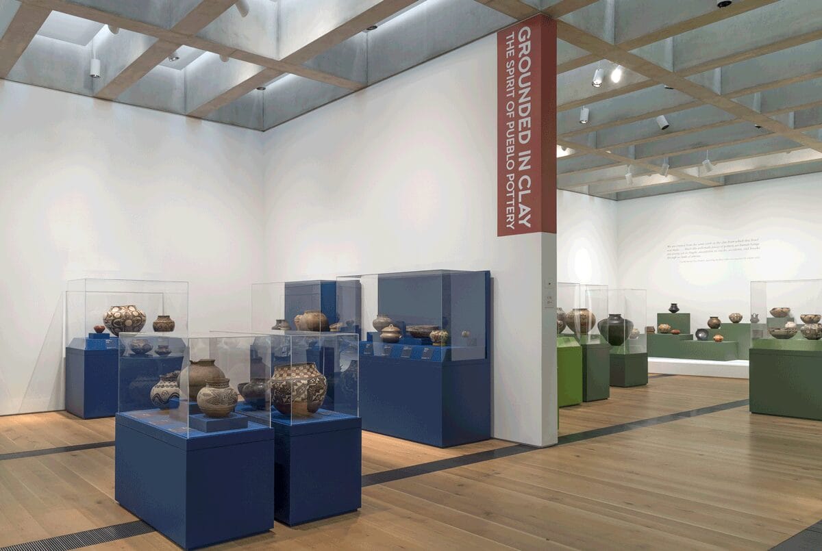 Multiple pieces of pottery in green and blue display cases on a gallery floor. A section of wall in the gallery has the exhibition name painted on it.