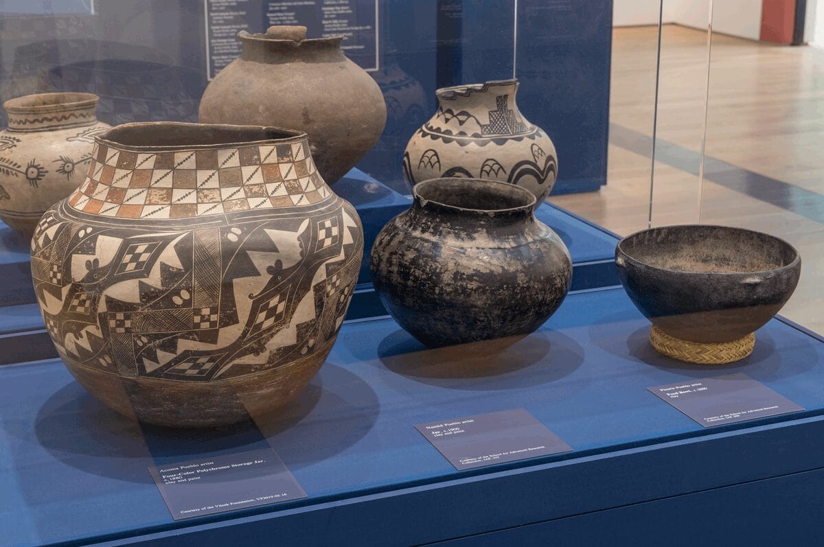 A dark blue display case with three clay pottery bowls placed within it; the pottery in the case behind is also visible.
