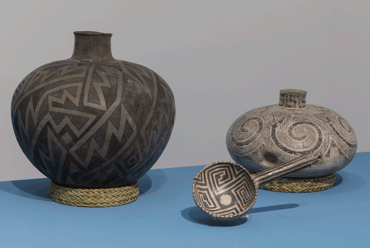 Three pottery pieces on view on a light blue pedestal. The two largest pieces are supported by pot rests made of yucca.