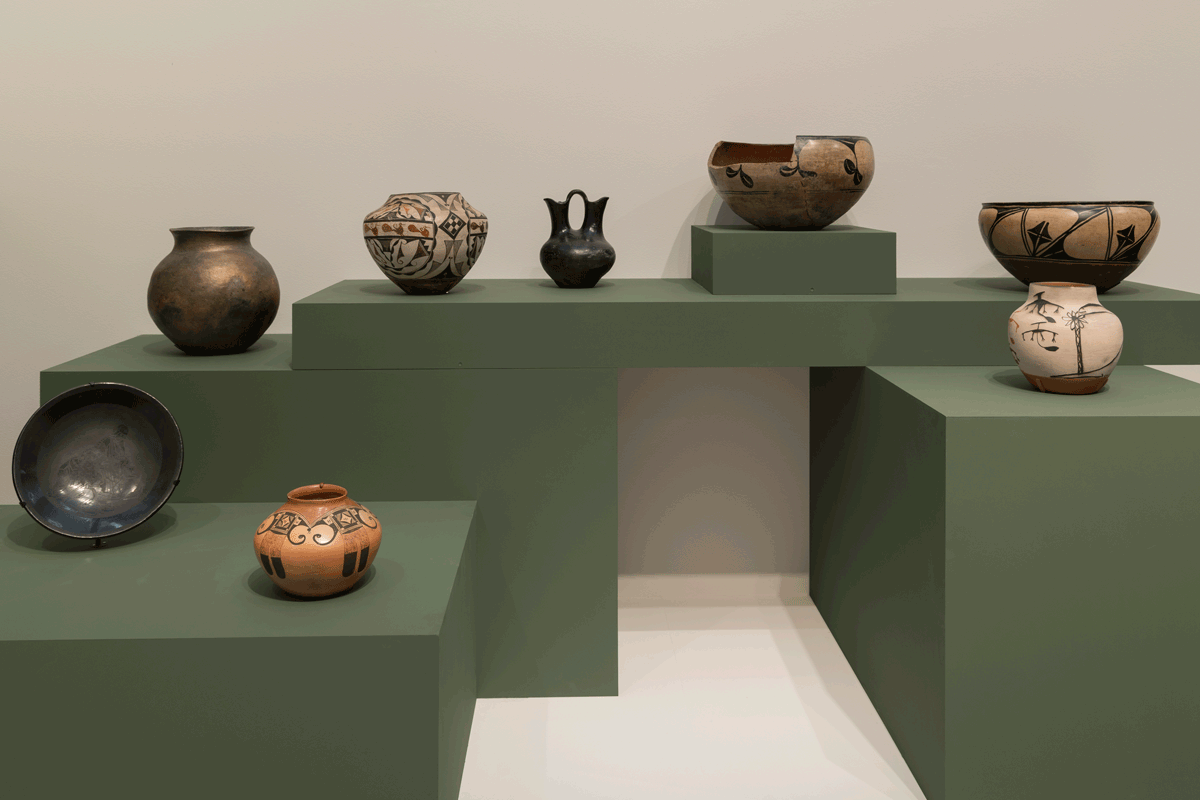 Eight clay pottery pieces decorated with pigments of beige, red, black, and orange displayed on green pedestals.