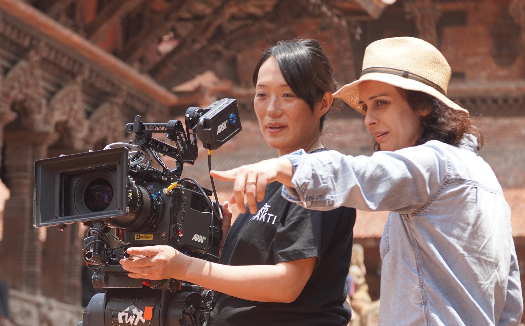Two filmmakers stand beside a professional ARRI camera on set, one adjusting the camera while the other points toward the scene in discussion.