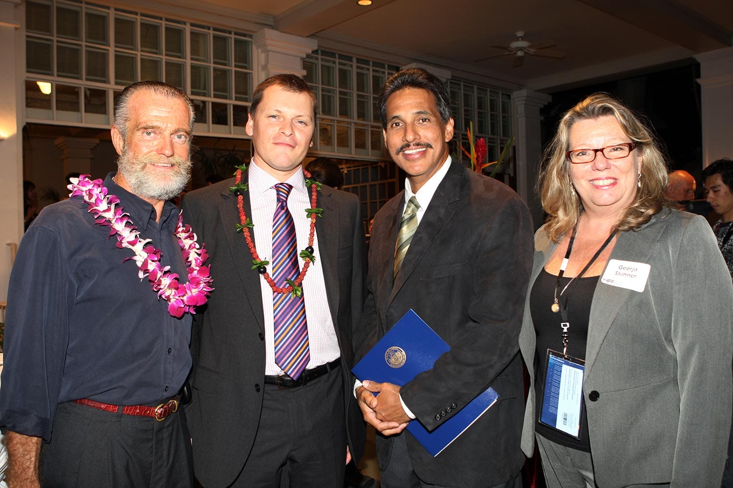 Four people stand together at an event, two wearing floral leis, all dressed in business attire and smiling for the photo.