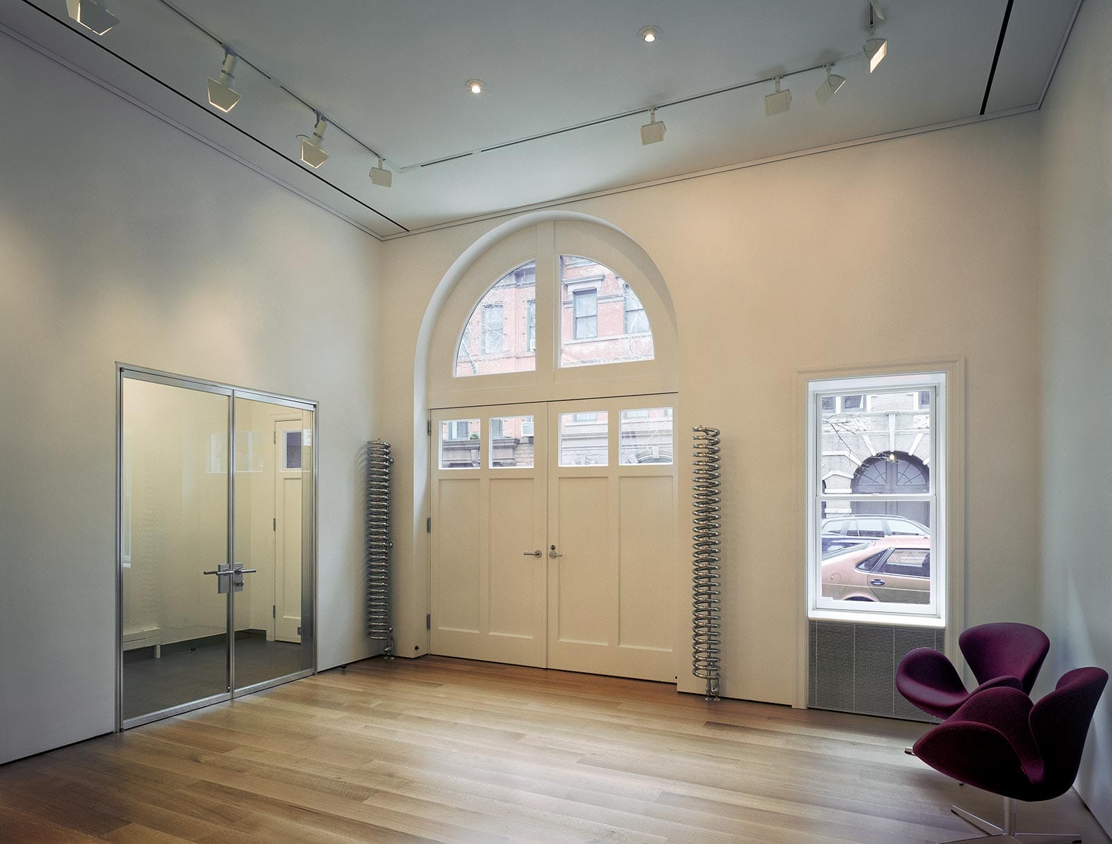 Empty gallery lobby with large carriage house doors, wood floors, and a maroon chair by a window.