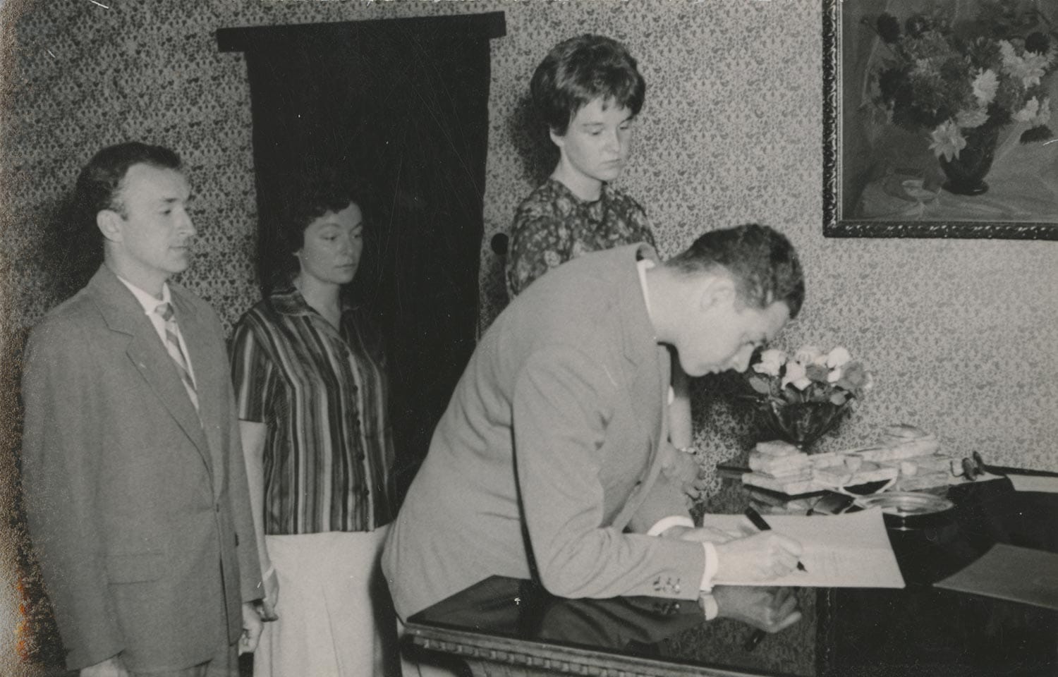 Jan Vilcek signs a document at a table while Marica Vilcek and two others stand behind him in a small, patterned room.